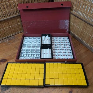 Op de afbeelding: Een complete Mahjong-set met een bordeauxrode koffer, met tegels met Chinese karakters en symbolen. De set bevat witte en gele tegels, samen met rekken om de spelstukken te organiseren. De koffer is open en onthult de georganiseerde spelcomponenten.