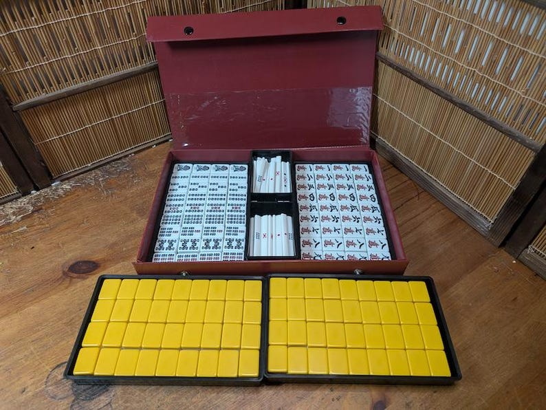 Op de afbeelding: Een complete Mahjong-set met een bordeauxrode koffer, met tegels met Chinese karakters en symbolen. De set bevat witte en gele tegels, samen met rekken om de spelstukken te organiseren. De koffer is open en onthult de georganiseerde spelcomponenten.