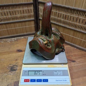 Op de afbeelding: Een gedetailleerd, antiek masker met een opvallend, omhoog wijzend kenmerk, geschilderd in tinten rood, groen en goud. Het masker staat op een digitale weegschaal, die 825g aangeeft. De achtergrond omvat een bamboe scherm en een houten oppervlak.
