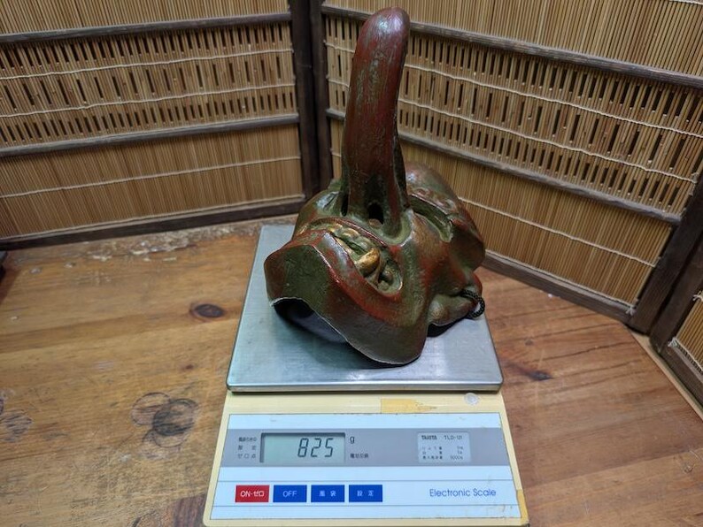 Op de afbeelding: Een gedetailleerd, antiek masker met een opvallend, omhoog wijzend kenmerk, geschilderd in tinten rood, groen en goud. Het masker staat op een digitale weegschaal, die 825g aangeeft. De achtergrond omvat een bamboe scherm en een houten oppervlak.