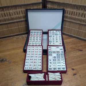May include: A vintage Mahjong set with ivory-coloured tiles featuring red, green, and blue symbols and characters. The set includes a black case, four tile racks, and a small red box with white sticks. The game is displayed on a wooden surface.