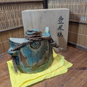 Puede incluir: Una botella de sake de cerámica con un diseño floral en tonos azules y verdes, atada con una cuerda marrón. Se encuentra sobre un paño amarillo, con una caja de madera detrás. La caja tiene caracteres japoneses.