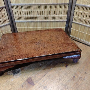 May include: A rectangular, wooden table with a glossy, speckled brown surface. The table has a dark brown, ornate base with four short legs. The background features a bamboo screen.