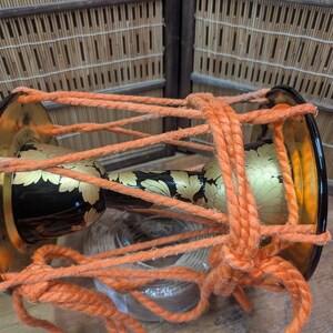 K&ouml;nnte beinhalten: Traditionelle japanische Trommel mit schwarzem Korpus und goldenen Blumenmustern. Sie ist mit dickem orangefarbenem Seil befestigt. Die Trommelenden sind gold und schwarz. Der Hintergrund zeigt Bambus und Holz.