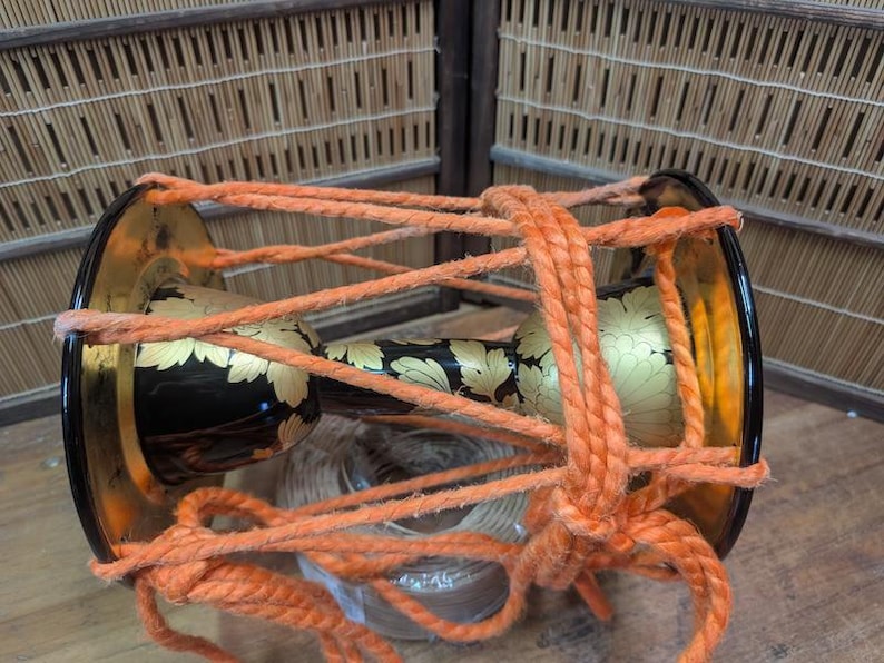 K&ouml;nnte beinhalten: Traditionelle japanische Trommel mit schwarzem Korpus und goldenen Blumenmustern. Sie ist mit dickem orangefarbenem Seil befestigt. Die Trommelenden sind gold und schwarz. Der Hintergrund zeigt Bambus und Holz.