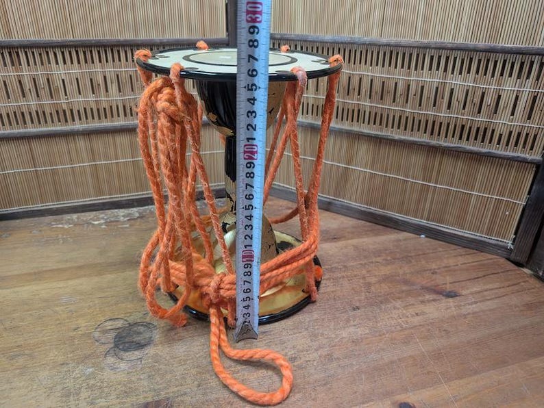 K&ouml;nnte beinhalten: Eine kleine, traditionelle japanische Trommel mit orangefarbenen Seilakzenten. Die Trommel ist schwarz und goldfarben, mit einer wei&szlig;en Oberseite. Ein Ma&szlig;band zeigt, dass die Trommel etwa 25 cm hoch ist.