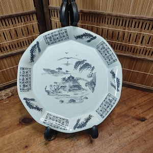Puede incluir: Plato de cerámica blanco con una escena de paisaje pintada a mano en tonos grises y azules. El plato tiene forma octogonal con patrones geométricos decorativos y detalles florales en los bordes. El plato se exhibe sobre un soporte negro.
