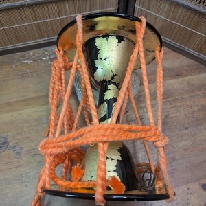 K&ouml;nnte beinhalten: Eine traditionelle japanische Sanduhr-f&ouml;rmige Trommel, oder Tsuzumi, mit einem schwarz lackierten Korpus, der mit floralen Goldblatt-Designs verziert ist. Die Trommel ist mit orangefarbenem Seil aufgeh&auml;ngt und hat einen goldenen Rand.
