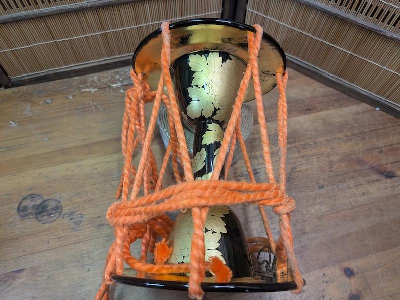 K&ouml;nnte beinhalten: Eine traditionelle japanische Sanduhr-f&ouml;rmige Trommel, oder Tsuzumi, mit einem schwarz lackierten Korpus, der mit floralen Goldblatt-Designs verziert ist. Die Trommel ist mit orangefarbenem Seil aufgeh&auml;ngt und hat einen goldenen Rand.