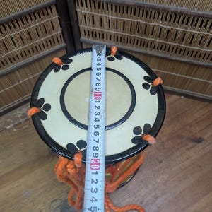 K&ouml;nnte beinhalten: Eine japanische Taiko-Trommel mit cremefarbenem Deckel und schwarzen floralen Designs. Die Trommel hat einen schwarzen Rand und ist mit orangefarbenem Garn verziert. Ein Lineal zeigt den Durchmesser der Trommel.