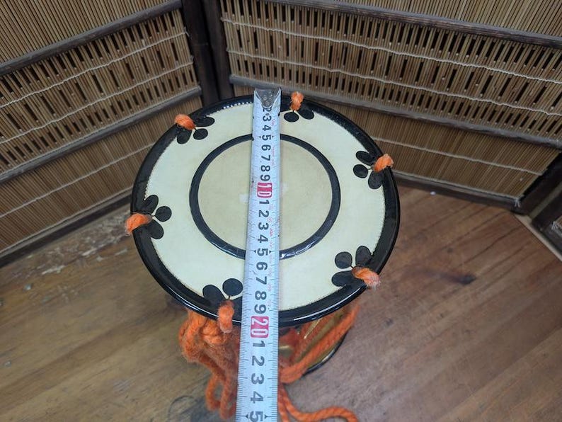 K&ouml;nnte beinhalten: Eine japanische Taiko-Trommel mit cremefarbenem Deckel und schwarzen floralen Designs. Die Trommel hat einen schwarzen Rand und ist mit orangefarbenem Garn verziert. Ein Lineal zeigt den Durchmesser der Trommel.