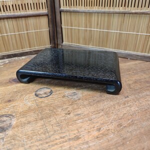 May include: A black, rectangular wooden display stand with rounded corners and a textured surface. The stand is supported by two curved feet. The stand is on a wooden surface.