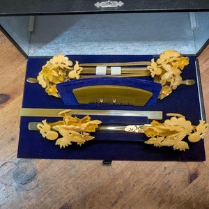 May include: A black box with a blue velvet interior contains a set of decorative hair accessories. The set includes gold-coloured hair sticks, combs, and floral decorations. The items are arranged neatly within the box.