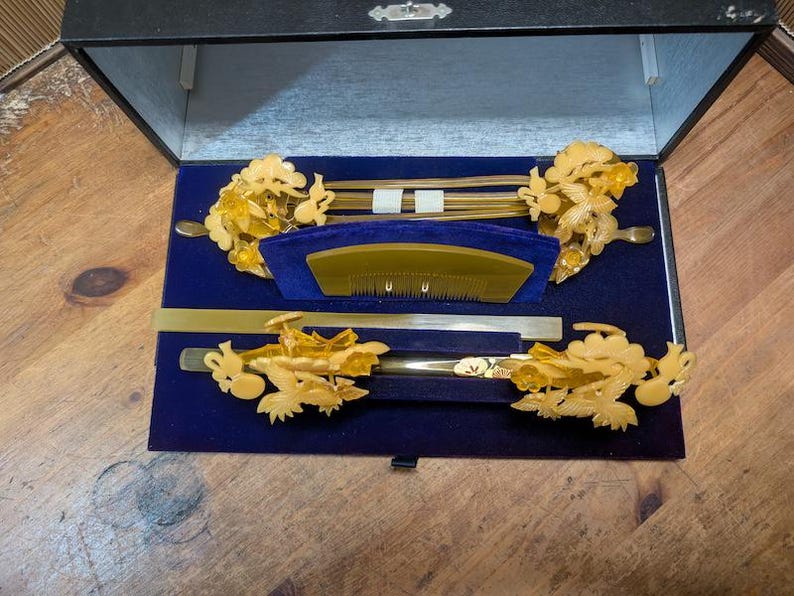 May include: A black box with a blue velvet interior contains a set of decorative hair accessories. The set includes gold-coloured hair sticks, combs, and floral decorations. The items are arranged neatly within the box.