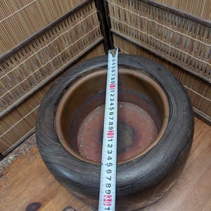 May include: A large, dark brown ceramic planter with a wide rim and textured surface. A measuring tape shows the planter's diameter. The planter is approximately 12 inches in diameter. The interior is a lighter brown color.