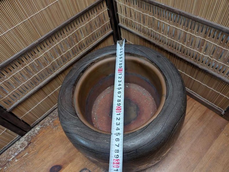 May include: A large, dark brown ceramic planter with a wide rim and textured surface. A measuring tape shows the planter's diameter. The planter is approximately 12 inches in diameter. The interior is a lighter brown color.