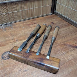 Pode incluir: Quatro formões de madeira com cabos de madeira e lâminas de metal são exibidos em um suporte de madeira. Os formões estão dispostos em fila, com as lâminas voltadas para a frente. O suporte de madeira é retangular e tem um acabamento castanho escuro.
