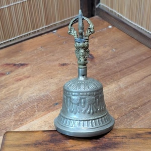 Puede incluir: Una campana de color plateado con un asa ornamentada con un diseño detallado y una carita. La campana tiene una base redondeada con un patrón decorativo. El asa está rematada con un elemento decorativo. La campana está sobre una superficie de madera.