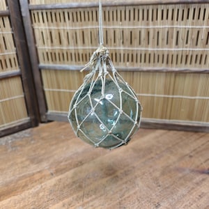 Op de afbeelding: Een heldere glazen visdrijver, opgehangen aan een geweven net en touw. De drijver is rond en heeft een lichte groene tint. De achtergrond heeft een houten structuur met een bamboe-achtig patroon.