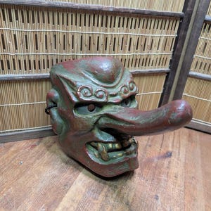 Op de afbeelding: Een vintage Japanse Tengu-masker, met een lange neus en een felle uitdrukking. Het masker is geschilderd in tinten rood en groen, met gouden accenten op de tanden. Het is gemaakt van een stevig materiaal en is een traditioneel Japans kunstwerk.