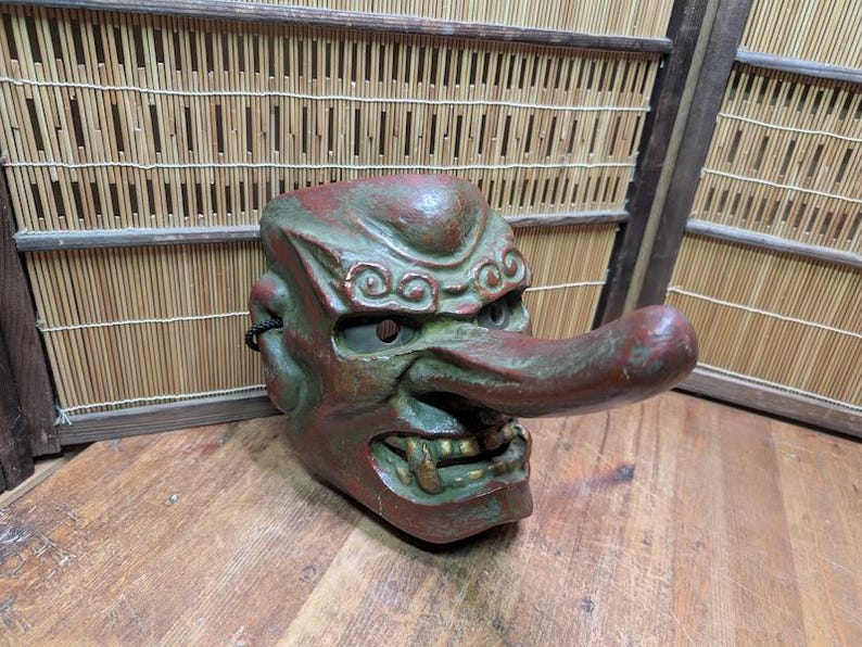 Op de afbeelding: Een vintage Japanse Tengu-masker, met een lange neus en een felle uitdrukking. Het masker is geschilderd in tinten rood en groen, met gouden accenten op de tanden. Het is gemaakt van een stevig materiaal en is een traditioneel Japans kunstwerk.