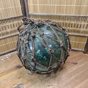 Puede incluir: Un flotador de pesca redondo de vidrio transparente con una red de cuerda marrón oscuro. El flotador tiene un ligero tinte verde y se encuentra sobre una superficie de madera. La red de cuerda está tejida alrededor de la esfera de vidrio.