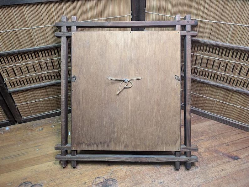 May include: A wooden framed square panel, likely the back of a mirror or artwork, with a natural wood grain finish. The frame is constructed of dark, rectangular wooden pieces joined at the corners. A natural fiber cord is tied to the center of the panel.