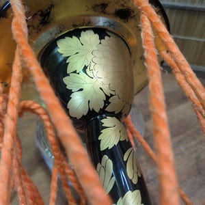 K&ouml;nnte beinhalten: Nahaufnahme einer traditionellen japanischen Trommel mit schwarzem Korpus und goldenem Blumendesign. Die Trommel hat einen goldenen Rand und ist mit orangefarbenem Seil verziert. Das Design der Trommel zeigt goldene Bl&auml;tter und eine gro&szlig;e Blume.