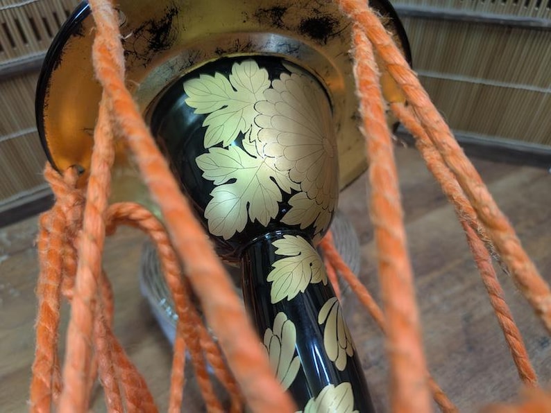 K&ouml;nnte beinhalten: Nahaufnahme einer traditionellen japanischen Trommel mit schwarzem Korpus und goldenem Blumendesign. Die Trommel hat einen goldenen Rand und ist mit orangefarbenem Seil verziert. Das Design der Trommel zeigt goldene Bl&auml;tter und eine gro&szlig;e Blume.