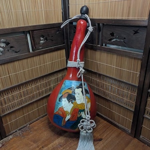 May include: A red gourd-shaped decorative bottle with a painted image of two people in traditional clothing. The bottle has a silver-colored cord and tassel. The background is a wooden structure with bamboo and carved panels.