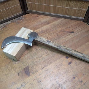 Puede incluir: Una hacha vintage con una hoja curva de color plateado y un mango de madera. El hacha descansa sobre un bloque de madera de color claro, colocado sobre una superficie de madera. El mango muestra signos de desgaste, lo que sugiere edad y uso.