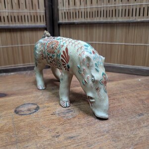 May include: A ceramic figurine of a hippopotamus with a cream-colored base and detailed floral patterns in red, teal, and green. The figurine is posed with its head lowered, and its legs are slightly bent.