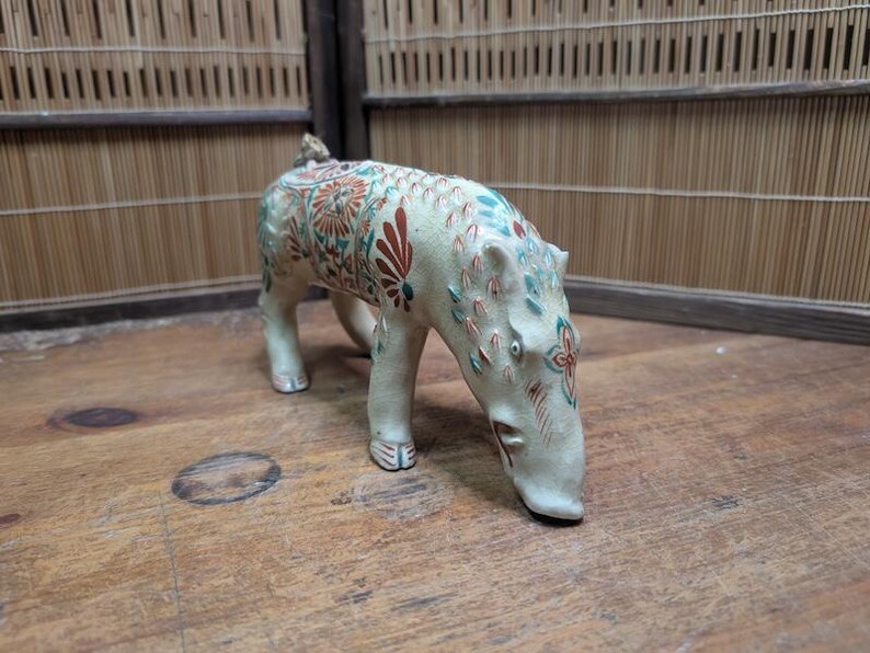 May include: A ceramic figurine of a hippopotamus with a cream-colored base and detailed floral patterns in red, teal, and green. The figurine is posed with its head lowered, and its legs are slightly bent.