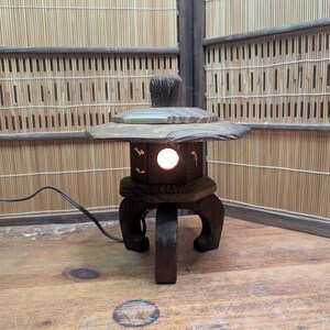 May include: A wooden Japanese-style lantern with a light inside. The lantern has a square base with curved legs and a sloped top. The lantern is dark brown and sits on a wooden surface. The background is a bamboo screen.