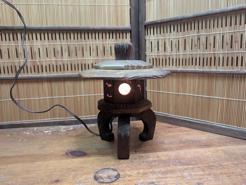 May include: A wooden Japanese-style lantern with a light inside. The lantern has a square base with curved legs and a sloped top. The lantern is dark brown and sits on a wooden surface. The background is a bamboo screen.