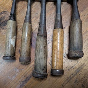 Puede incluir: Primer plano de cinco cinceles de madera antiguos. Cada cincel tiene una hoja de metal y un mango de madera, que muestran signos de desgaste y envejecimiento. Los mangos var&iacute;an en color, desde marr&oacute;n claro hasta gris oscuro. Los cinceles est&aacute;n dispuestos verticalmente.