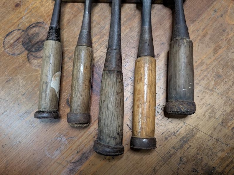 Puede incluir: Primer plano de cinco cinceles de madera antiguos. Cada cincel tiene una hoja de metal y un mango de madera, que muestran signos de desgaste y envejecimiento. Los mangos var&iacute;an en color, desde marr&oacute;n claro hasta gris oscuro. Los cinceles est&aacute;n dispuestos verticalmente.