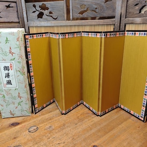 May include: A four-panel folding screen with a solid gold-colored surface and a decorative border featuring a repeating geometric pattern in red, white, and blue. The screen is accompanied by a rectangular storage box with a crane print and text.