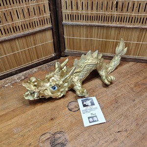 May include: A golden dragon figurine with blue eyes and intricate detailing. The dragon is posed on a wooden surface, with a small white card nearby. The dragon's scales and features are clearly defined, showcasing a mythical creature.
