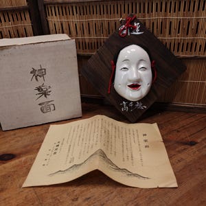 May include: A white ceramic mask with black hair and red lips mounted on a dark brown diamond-shaped wooden plaque with white Japanese calligraphy. The set includes a white cardboard box with black text and a folded paper document with text.