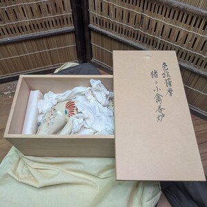 May include: A wooden box with a ceramic pig figurine inside, cushioned by white fabric. The figurine is cream-colored with red and blue accents. The box is open, with a separate lid featuring Japanese text.