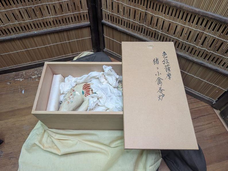 May include: A wooden box with a ceramic pig figurine inside, cushioned by white fabric. The figurine is cream-colored with red and blue accents. The box is open, with a separate lid featuring Japanese text.
