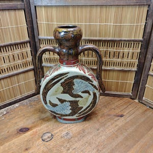 May include: A round ceramic vessel with two arched handles and a narrow neck. The body features a cream-colored base with dark brown and light blue abstract leaf-like patterns. A red band encircles the neck, and the top rim is dark brown.