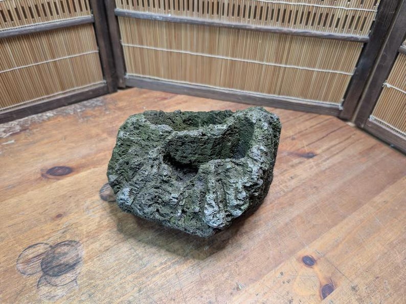 Puede incluir: Una maceta cuadrada, texturizada, similar a la piedra, con un exterior rugoso y musgoso. La maceta tiene una cavidad central y es principalmente gris y verde. Se encuentra sobre una superficie de madera, lo que sugiere una est&eacute;tica natural o r&uacute;stica.