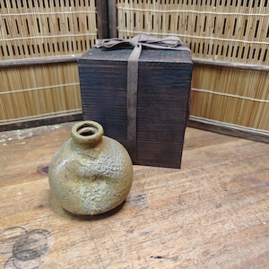 May include: A small, round, textured ceramic vase with a narrow neck sits on a wooden surface next to a dark wooden box tied with a ribbon. The vase has a mottled, earthy color, and the box is rectangular.