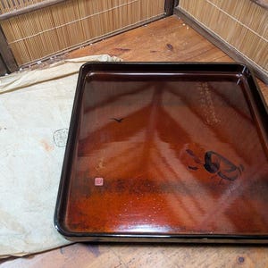 以下が含まれることがあります： 光沢のある赤茶色の仕上げが施された、長方形の漆塗りの木製トレイ。トレイは黒い縁取りと、鳥やその他の芸術的な詳細を含む装飾的な要素を備えています。トレイの横には紙が置かれています。