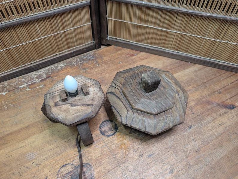 May include: A rustic wooden lamp with a unique, geometric design. The lamp features a light bulb and a separate wooden base. The wood has a natural, weathered appearance, adding to its charm. The lamp is designed to sit on a table or desk.