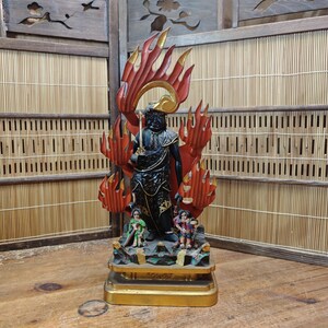 May include: A detailed statue of a figure in black robes, holding a sword, with red and gold flames behind it. Two smaller figures stand at the base of the statue. The statue is on a gold base, set against a bamboo screen.