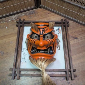 May include: An orange and black Japanese demon mask with a fierce expression, framed in a wooden border. The mask features detailed facial features and a long, light-colored hairpiece. The mask is mounted on a white background with Japanese calligraphy.