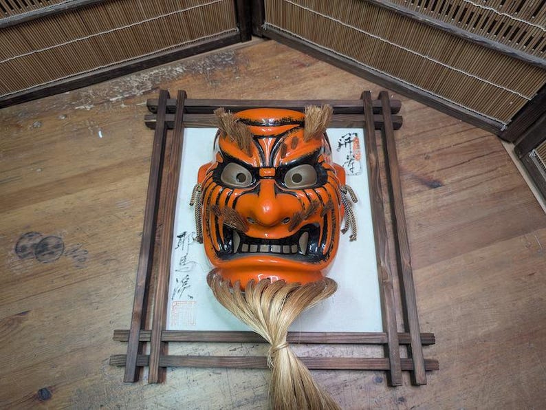 May include: An orange and black Japanese demon mask with a fierce expression, framed in a wooden border. The mask features detailed facial features and a long, light-colored hairpiece. The mask is mounted on a white background with Japanese calligraphy.